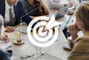 Geschäftstreffen an einem Tisch mit Laptops und Notizblöcken. Die Leute diskutieren. Ein transparentes Zielsymbol mit einem Pfeil ist darübergelegt und symbolisiert Fokus oder Zielsetzung.