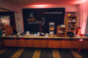 Ein Ausstellungstisch auf einer Veranstaltung mit verschiedenen Drucksachen und Werbeartikeln. Auf einem großen Schild steht: „Ohne Anfassen geht’s nicht!“ Der Stand trägt den Schriftzug „Onlineprinters“. In Holzkisten liegen Flyer und Karten mit verschiedenen Preisen.