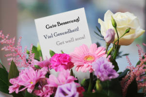 Ein Strauß rosa und lila Blumen mit einer weißen Rose. Auf einer Karte zwischen den Blumen steht in schwarzer Schrift: „Gute Besserung! Viel Gesundheit! Gute Besserung!“.