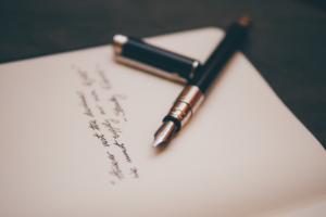 Ein Füllfederhalter mit schwarzem Schaft und silbernen Akzenten liegt auf einem offenen Notizbuch. Die Kappe des Stifts liegt daneben. Auf den cremefarbenen Seiten des Notizbuchs ist handgeschriebener Text in schwarzer Tinte zu sehen. Der Hintergrund ist unscharf.