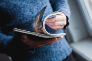 Eine Person in einem blauen Pullover blättert in der Nähe eines sonnenbeschienenen Fensters in einem Kundenmagazin.
