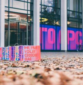 Bunte Goodie Bags reihen sich auf dem Boden aneinander, auf einer steht stolz: „Hi, ich bin deine Goodie Bag.“ Im Hintergrund bildet ein modernes Gebäude mit großen Glasfenstern und leuchtenden Neonschildern die Kulisse, während überall Laub verstreut liegt. Perfekt für jede Give-away-Messe!.