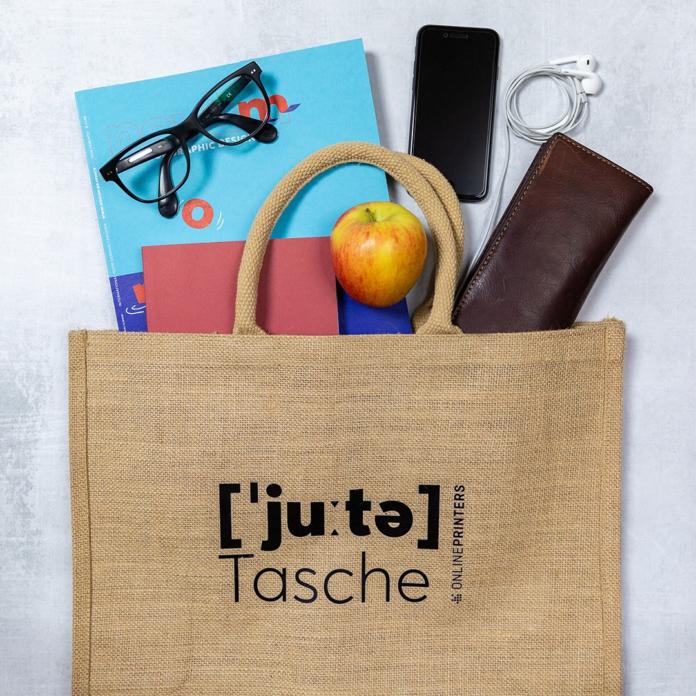 Eine Jutetasche mit roten Henkeln, bestückt mit Give-away-Materialien der Messe, enthält ein blaues Notizbuch, eine Brille, einen Apfel, Ohrhörer, ein schwarzes Smartphone und eine braune Geldbörse. Stilvoll platziert auf einem hellgrauen Untergrund.