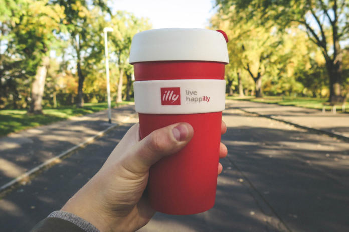 Eine Hand hält einen roten Mehrweg-Kaffeebecher, perfekt zum Verschenken, mit weißem Deckel und dem Logo „illy live happilly“. Im Hintergrund erstreckt sich unter dem klaren Himmel ein von Bäumen gesäumter Park mit einem gepflasterten Weg.