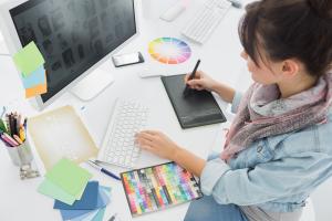 Eine Frau sitzt an einem Schreibtisch und benutzt ein Zeichentablett, während sie auf einen Computerbildschirm blickt. Der Schreibtisch ist übersät mit Farbmustern, einem Farbrad, Notizblöcken, einem Smartphone und bunten Papieren. Sie trägt einen Schal und eine Jeansjacke.