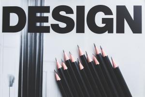 Eine Gruppe schwarzer Bleistifte ist kunstvoll auf einer Fläche angeordnet. Darüber steht in fetten, schwarzen Buchstaben das Wort „DESIGN“. Im Hintergrund ist ein verschwommenes Bild vertikaler Linien zu sehen, die an Vorhänge erinnern.