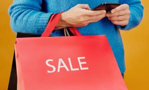Eine Person in einem blauen Pullover hält eine rote Einkaufstüte mit der Aufschrift „SALE“ und bedient gleichzeitig ein Smartphone. Der Hintergrund ist in einem warmen, gelblichen Farbton gehalten.
