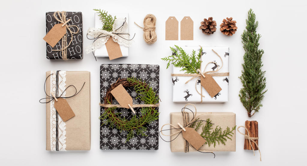 Eine Auswahl an Weihnachtsgeschenken für Kunden, verpackt in festliches Papier mit verschiedenen Motiven, mit Bindfaden zusammengebunden und mit Grünpflanzen, Tannenzapfen und Zimtstangen geschmückt. Leere Geschenkanhänger liegen daneben und sorgen für eine rustikale Weihnachtsdekoration auf weißem Hintergrund.