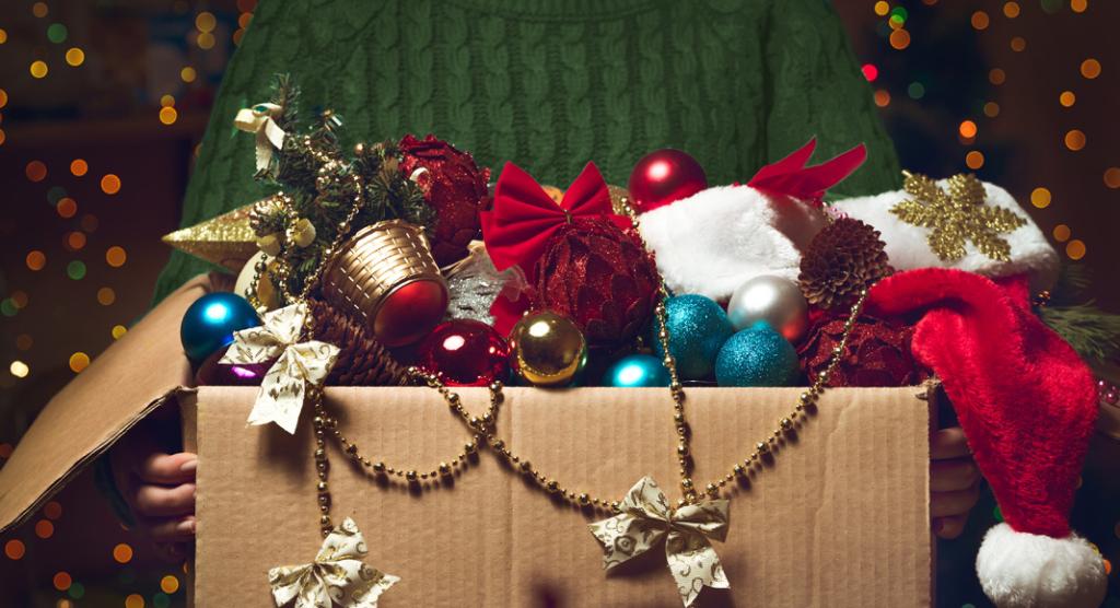 Eine Person in einem grünen Pullover hält eine Schachtel voller Weihnachtsdekorationen, Ornamenten, einer Weihnachtsmütze, Tannenzapfen und Goldperlen – eine perfekte Szene für die Auswahl von Weihnachtsgeschenken für Kunden. Festliche Lichter erzeugen einen warmen, verschwommenen Hintergrund.