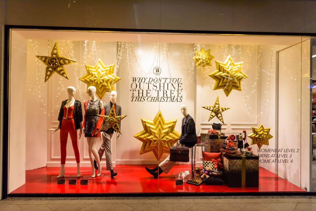 Schaufensterdekoration mit glitzernden goldenen Sternen, Schaufensterpuppen in festlicher Kleidung und der Botschaft „Warum überstrahlen Sie dieses Weihnachten nicht den Weihnachtsbaum?“. Rote Deko-Akzente und verschiedene Geschenkboxen runden das Weihnachtsthema ab.