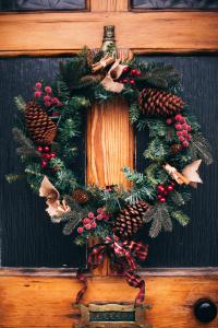 Ein festlicher Weihnachtskranz mit Tannenzapfen, roten Beeren und Bandverzierungen hängt an einer Holztür mit Briefschlitz. Der Kranz besteht aus immergrünen Zweigen und ist mit kleinen Jute-Akzenten verziert.