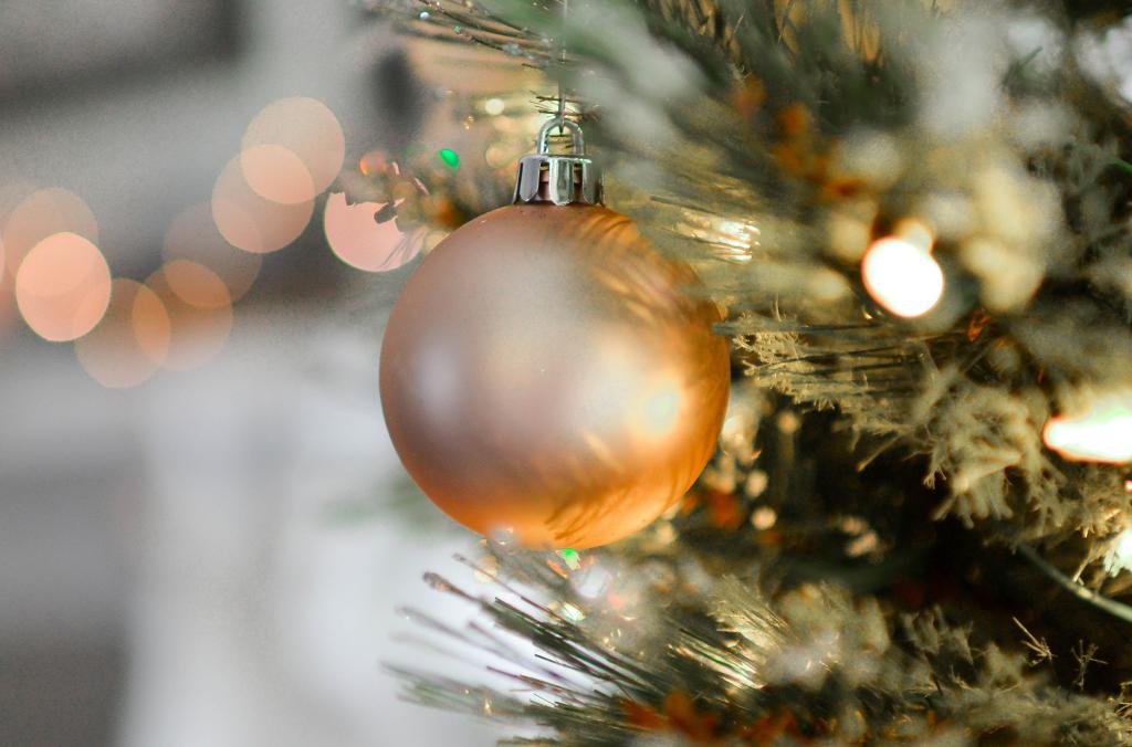 Eine Nahaufnahme eines orangefarbenen Weihnachtsornaments mit grünen Nadeln, das an einem Baum hängt. Weiße und warme gelbe Lichter erzeugen ein gemütliches Leuchten. Im Hintergrund erscheinen verschwommene runde Lichter, die die festliche Atmosphäre verstärken.