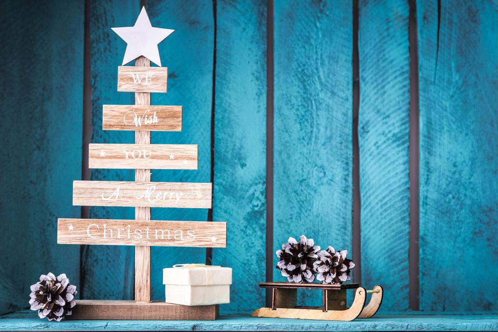 Ein hölzerner Christbaumschmuck mit dem Text „Wir wünschen Ihnen frohe Weihnachten“ steht neben einer kleinen Geschenkbox und einem Schlitten mit Tannenzapfen. Der Hintergrund ist eine strukturierte blaue Wand.