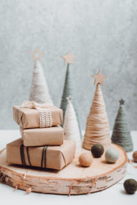 Eine festliche Szene mit kleinen, mit Garn umwickelten Zapfenbäumen mit Sternen an der Spitze, einigen Filzbällen und drei in braunes Papier und Schnur eingewickelten Geschenkboxen – perfekte Weihnachtsgeschenke für Kunden – alles präsentiert auf einer Holzscheibe vor einem hellgrauen Hintergrund.