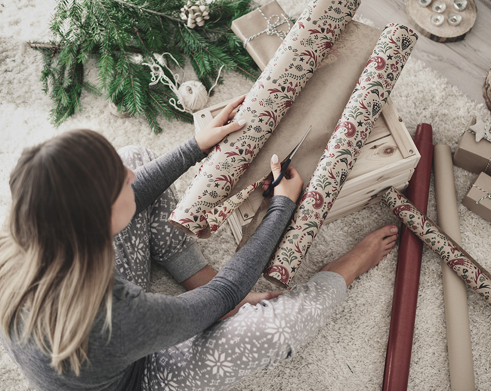 Eine Person sitzt auf einem Teppichboden und schneidet mit einer Schere festlich gemustertes Geschenkpapier zu, um Weihnachtsgeschenke für Kunden vorzubereiten. Umgeben von mehreren Geschenkpapierrollen und verpackten Geschenken sorgen die grünen Tannenzweige in der Nähe für festliche Stimmung.