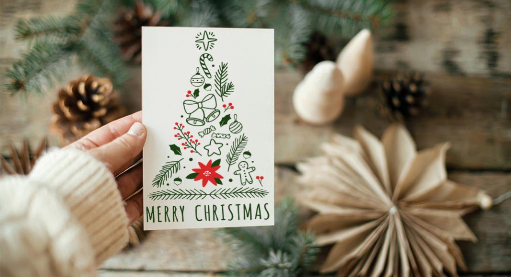 Eine Hand hält eine festliche Karte mit einem Weihnachtsbaum, der aus Weihnachtssymbolen wie Weihnachtsschmuck, Zuckerstangen und einem Lebkuchenmann gestaltet ist. Unten steht der elegante Schriftzug „Frohe Weihnachten“. Tannenzapfen und Tannenzweige zieren die Holzoberfläche dahinter – perfekt für geschäftliche Weihnachtsgrüße.