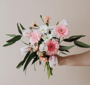Eine Hand hält sanft einen Strauß rosa Gerbera-Gänseblümchen, weiße und rosa Tulpen und zarte kleine weiße und pfirsichfarbene Blüten, akzentuiert mit grünen Blättern vor einem hellbeigen Hintergrund – perfekt für inspirierende Gutschein-Textvorlagen.