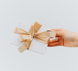 Eine mit zwei Ringen geschmückte Hand präsentiert eine weiße Geschenkbox mit kleinen Sternen und einer glänzenden Goldschleife, perfekt für Gutschein-Textvorlagen. Der Hintergrund ist schlicht und sanft weiß.