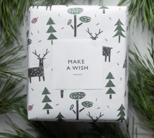 Ein Geschenk in weißem Papier, verziert mit grünen Bäumen und schwarzen Rentieren, perfekt als Weihnachtsgeschenk für Kunden. Die Schachtel ist von Tannenzweigen umgeben. Auf dem weißen quadratischen Etikett steht „WÜNSCH DIR WAS“.
