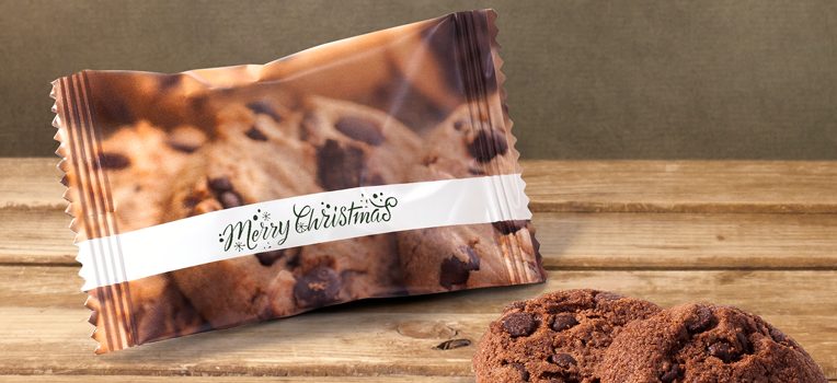 Eine Packung Chocolate Chip Cookies mit der Aufschrift „Frohe Weihnachten“ liegt auf einem Holztisch – perfekte Weihnachtsgeschenke für Kunden. Zwei Kekse liegen neben der Packung und zeigen ihre köstliche Textur in Nahaufnahme.