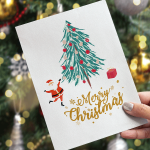 Eine Hand hält eine „Frohe Weihnachten“-Karte mit einem festlichen Motiv: ein geschmückter Weihnachtsbaum, eine kleine Weihnachtsmannfigur und ein Geschenk. Verschwommene Lichter und Ornamente schaffen einen warmen Hintergrund – perfekt für alle, die Weihnachtsgeschenke für Kunden suchen.