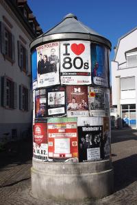 Ein zylindrischer Kiosk in einer Straße ist mit bunten Plakaten bedeckt, darunter ein auffälliges „I Love 80s“-Schild. In der Nähe sind Gebäude und ein Fahrrad zu sehen, was auf eine städtische Umgebung hindeutet.