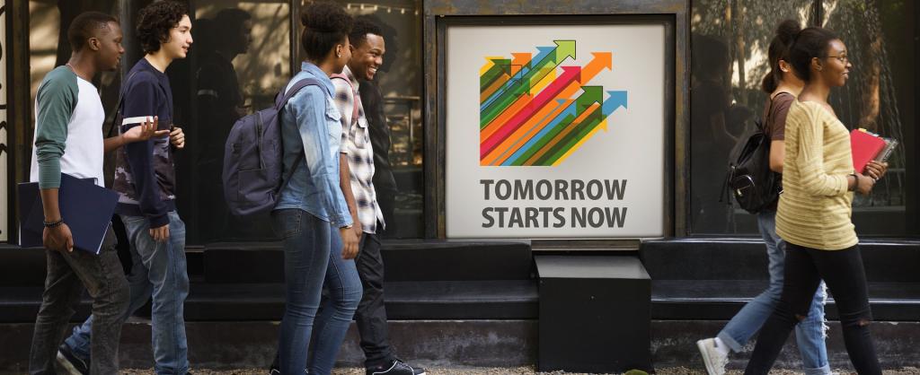 Eine bunt gemischte Gruppe lächelnder Studierender mit Rucksäcken geht an einem Gebäude vorbei. Ein großes Plakat am Fenster zeigt bunte Pfeile nach oben mit dem Text „Tomorrow Starts Now“. In den Fenstern spiegeln sich Bäume.